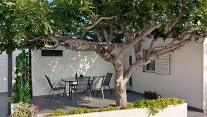 Un área de estar al aire libre con una mesa y sillas bajo un árbol, rodeada de paredes blancas y una ventana con contraventanas.
