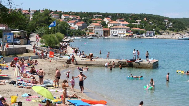 Une plage où des gens se prélassent au soleil et se baignent, une jetée avec des bateaux, et des maisons en arrière-plan.