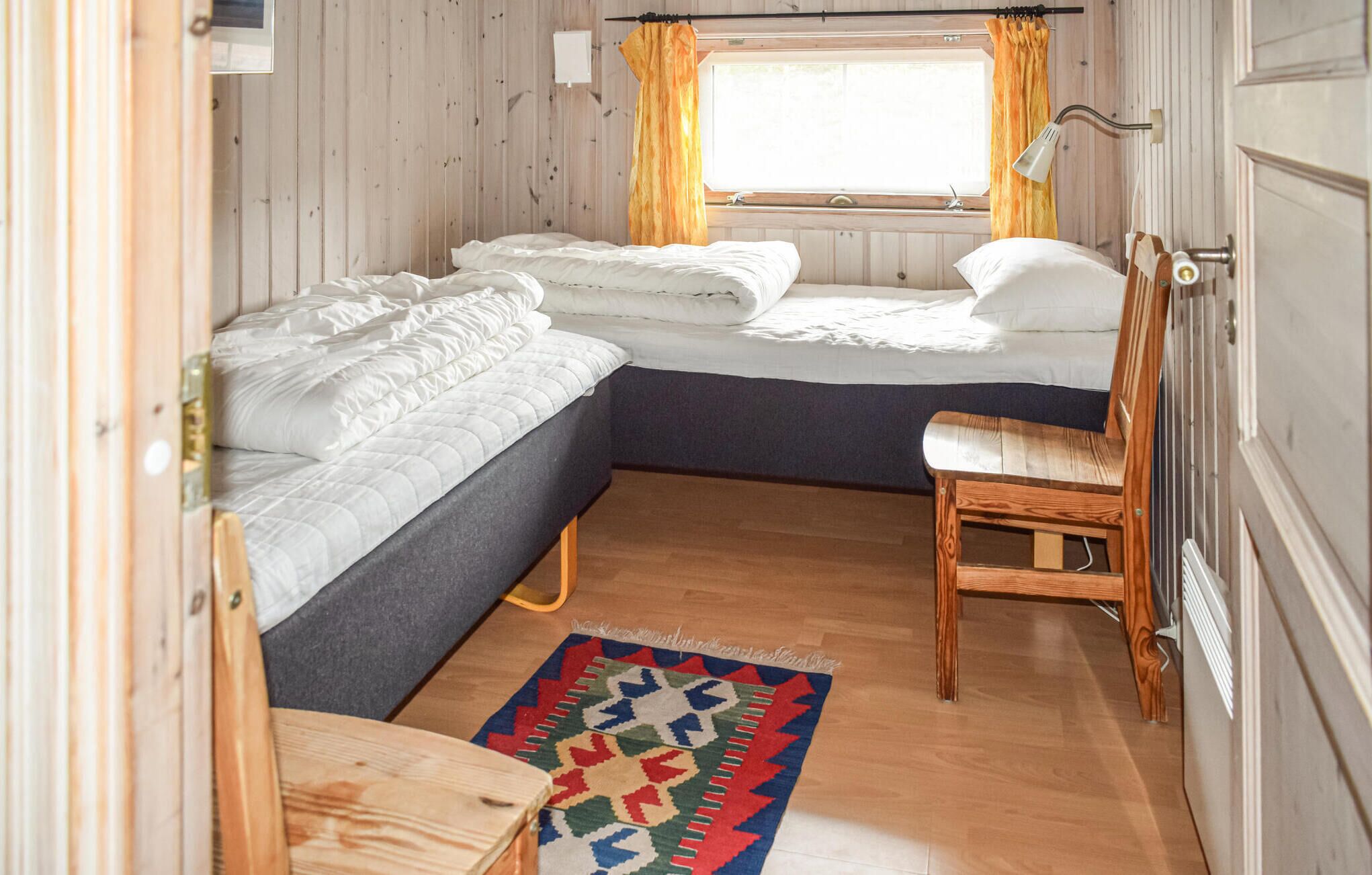 A compact bedroom with two beds, a small table, a chair, a window with curtains, and a radiator.
