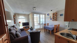 Ein Wohnzimmer mit einer Couch, einem Couchtisch und einem Fernseher. Es gibt einen Essbereich mit Tisch und Stühlen. Der Raum hat Holzböden und eine Küchenzeile mit Spüle und Schränken.