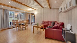 A cozy living room with a wooden floor, a red leather sofa, a wooden dining table with chairs, and a large window providing a view of the outside. - 3 bedroom beach front home in Højby (Højby)