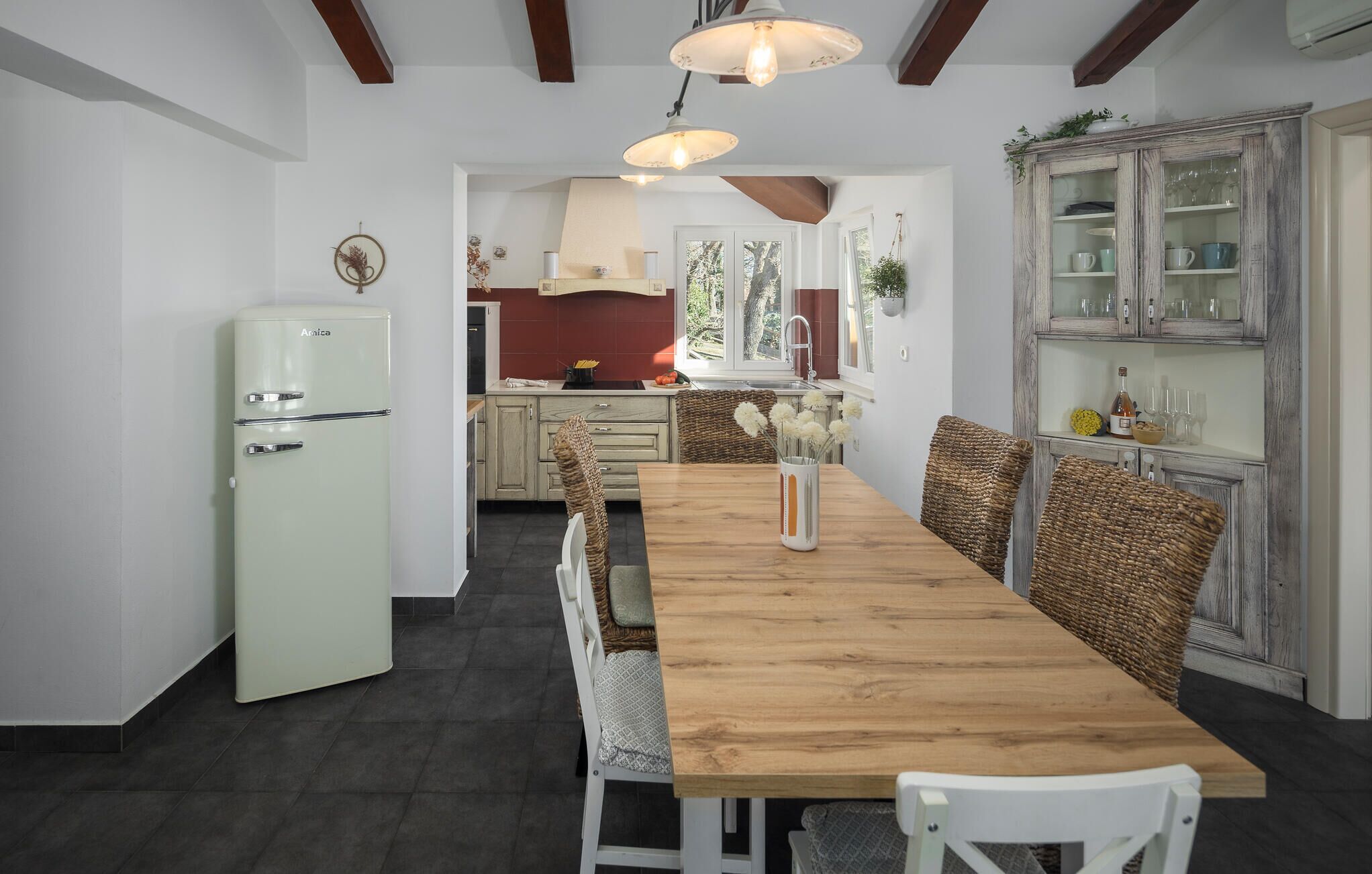 Una cocina con una mesa de madera, sillas de mimbre, un refrigerador antiguo y un armario con puertas de vidrio.