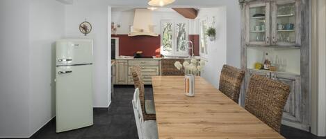 Una cocina con una mesa de madera, sillas de mimbre, un refrigerador antiguo y un armario con puertas de vidrio.