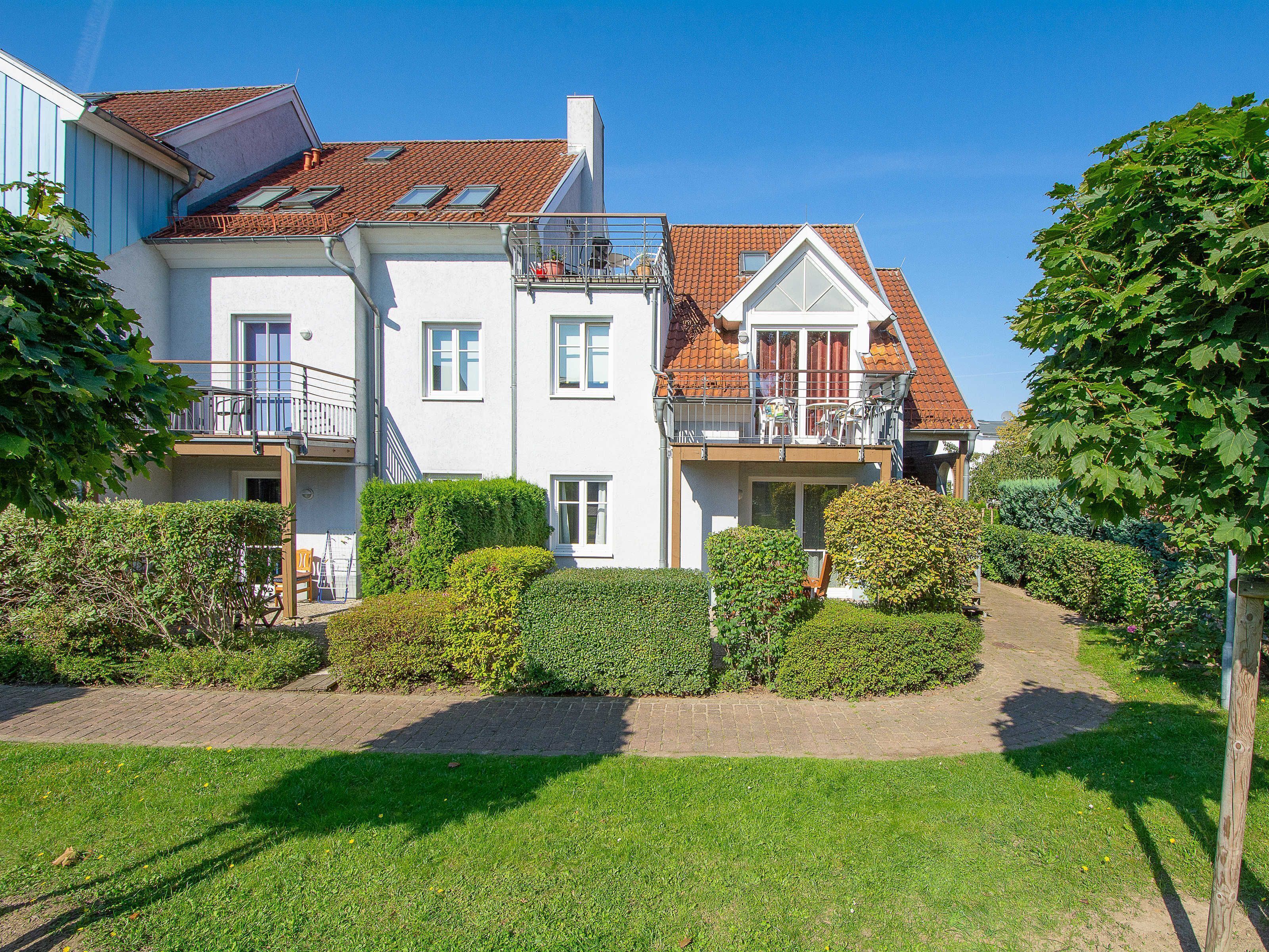 A two-story house with a red-tiled roof, a balcony with a railing, and a well-maintained garden with hedges and a paved pathway.