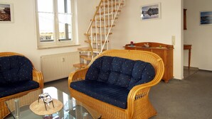 Een woonkamer met rieten meubels, een glazen salontafel, een houten trap en uitzicht op buiten.