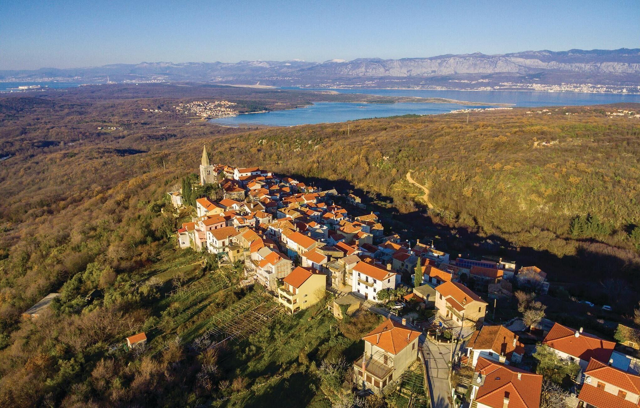 An aerial view of a hillside village with terraced houses, a church, and a lake in the background.