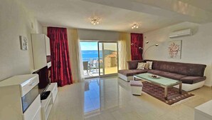 Un salon moderne avec un canapé, une table basse et une vue sur la mer, visible à travers une porte-fenêtre coulissante.