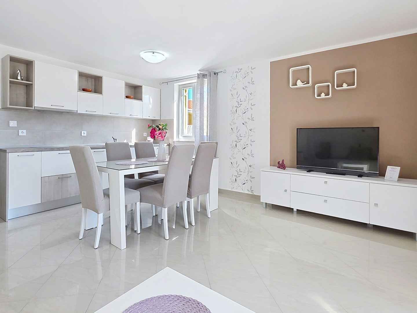 Une cuisine moderne avec des meubles blancs, un coin repas comprenant une table et des chaises, un téléviseur posé sur un meuble blanc, et un mur décoratif orné de tableaux encadrés.