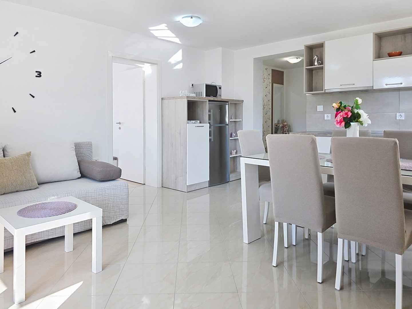 A modern living room with a sofa, a dining table with chairs, a kitchen area with a refrigerator, and a white door.
