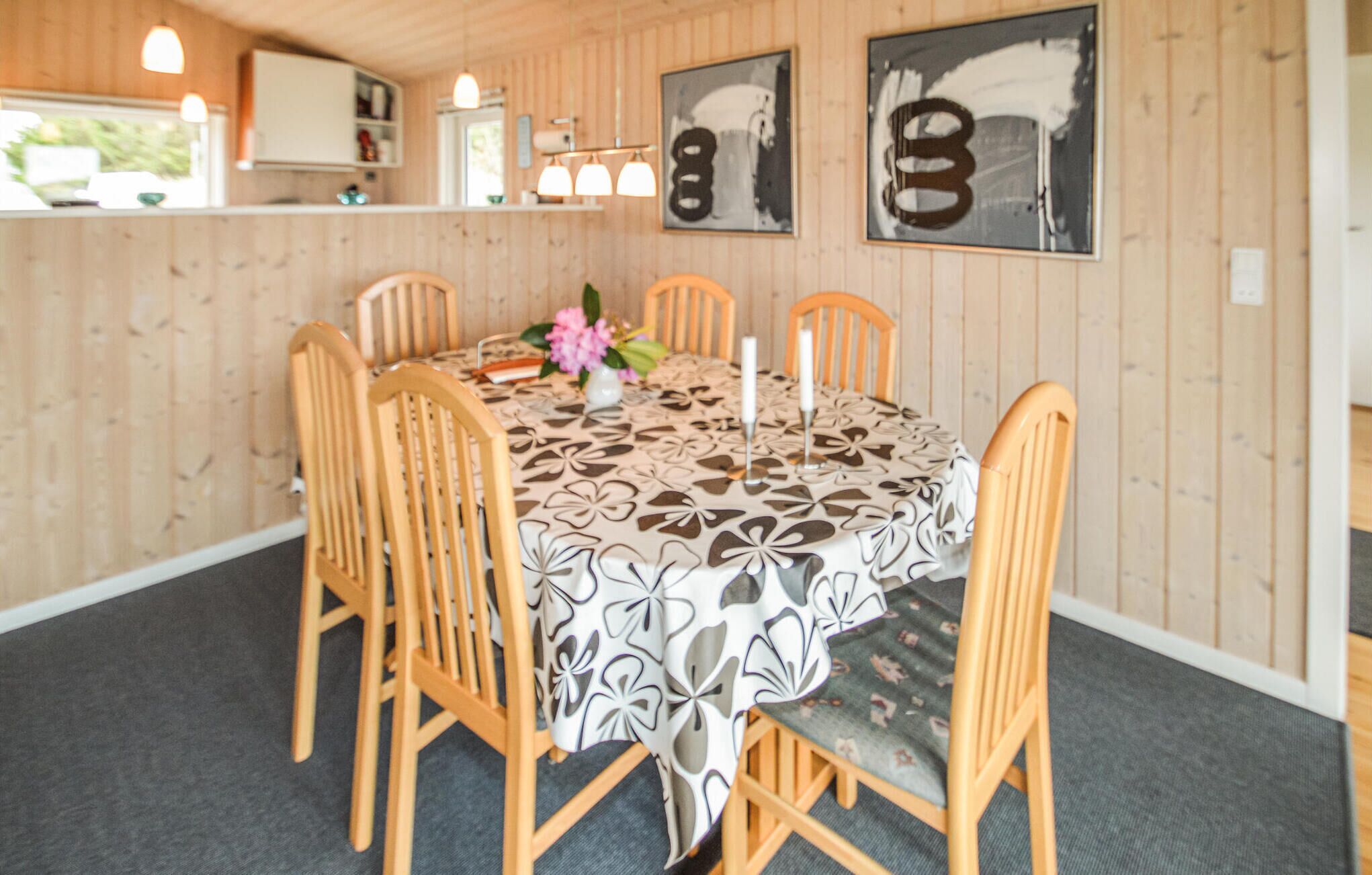 Ein Essbereich mit einem Tisch, gedeckt für vier Personen, umgeben von Holzsesseln. Auf dem Tisch liegt eine Tischdecke mit Muster, eine Vase mit Blumen und Kerzen. Der Raum hat Holzwände und im Hintergrund ist eine Küche zu sehen.