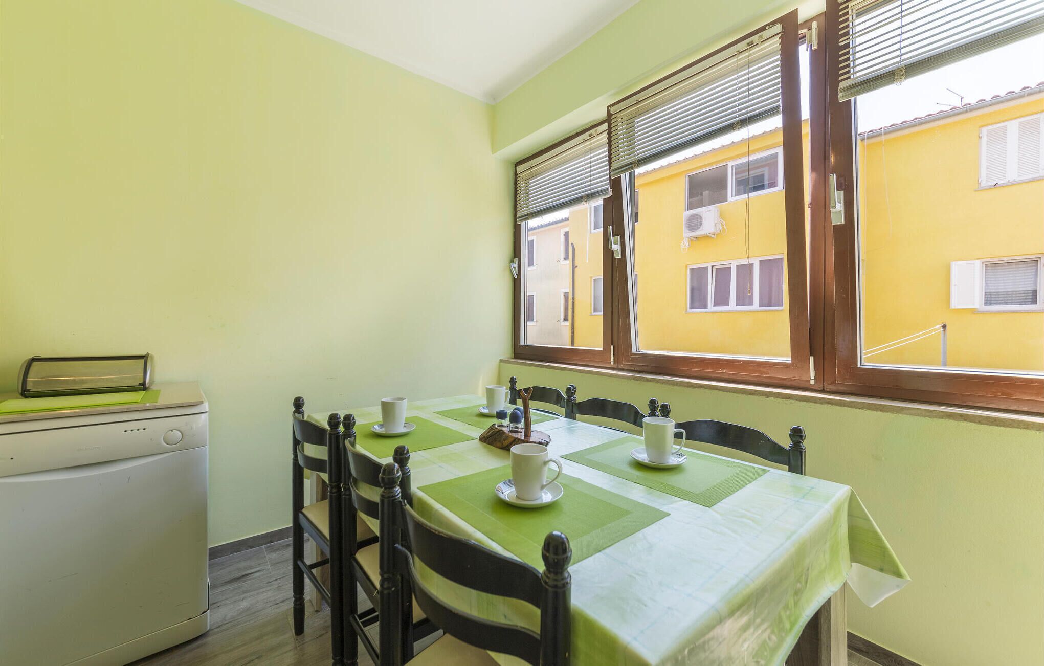 A dining area with a table set for two, a small kitchen area with a washing machine, and a window with blinds.