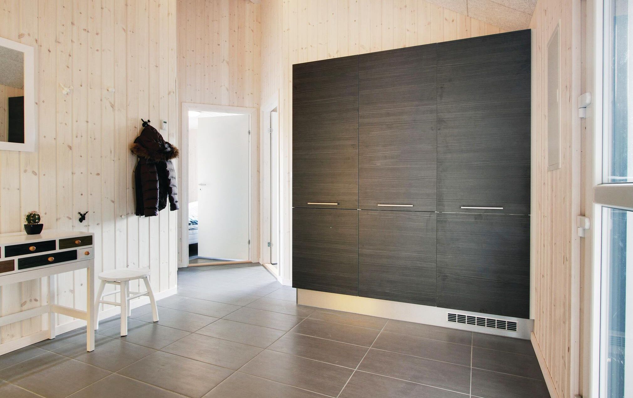 Una habitación moderna con piso de madera, un escritorio blanco con cajones, un perchero con abrigo y un amplio armario oscuro de dos puertas.