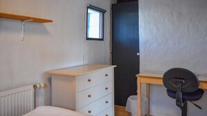 Een slaapkamer met een bed, een witte ladekast, een houten bureau met stoel, een radiator, een raam en een deur.