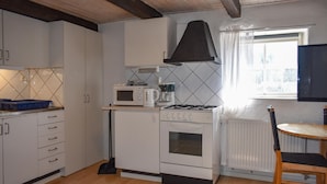 Een kleine keuken met witte kastjes, een magnetron, een fornuis en een koffiezetapparaat. Er is een raam met gordijnen en een radiator.