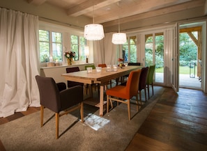 Un coin repas avec une table en bois, des chaises et un tapis. On y trouve des fenêtres avec des rideaux, un vase rempli de fleurs et une vue sur la verdure à l’extérieur.