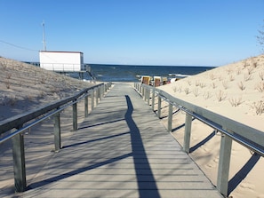 Une promenade en bois mène à une plage où se trouve un bâtiment blanc et un poste de surveillance des sauveteurs.
