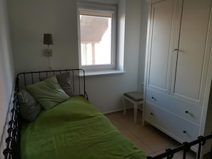 Un lit simple avec une couvre-lit verte, une lampe de chevet, une fenêtre, un petit tabouret et une armoire.