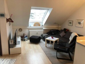 En mysig lägenhet på en vindsvåning med ett sluttande tak, en liten soffa, ett soffbord, ett element och ett fönster med utsikt över träd.
