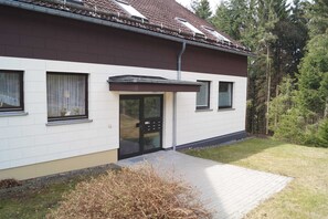 Ett modernt hus i två våningar med vit fasad och brunt tak, en ytterdörr och en liten trädgård framför huset.