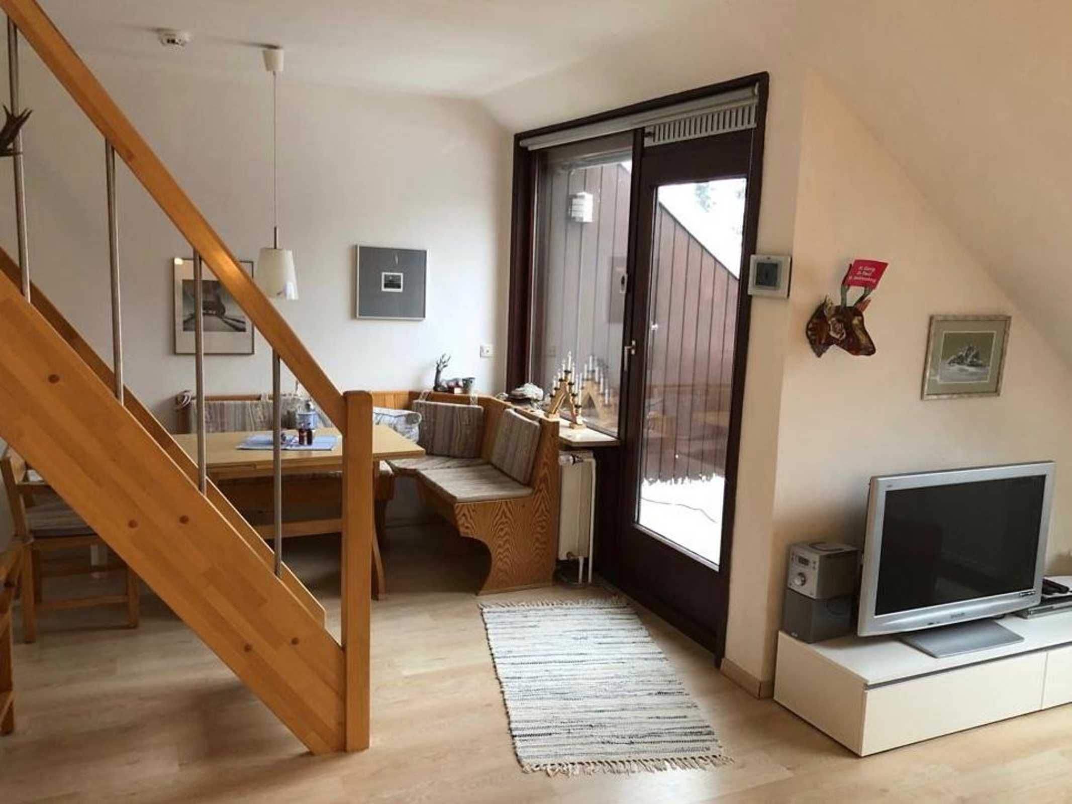 Uma sala de estar moderna com escada de madeira, área de jantar, televisão e uma grande janela.