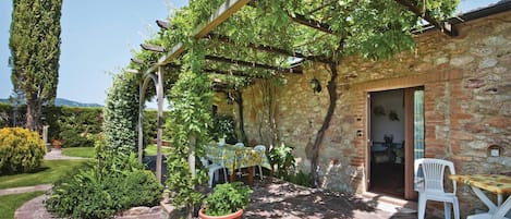 En bygning i sten med et pergola overdækket med grønt, udendørs siddepladser og en stensti.