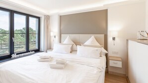 A hotel room with a large bed, a headboard with two light fixtures, a nightstand with a lamp, and a view of trees through the window.