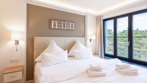 A hotel room with a large bed, white bedding, a headboard, two bedside lamps, a nightstand, a wooden side table, and a window with a view of trees.