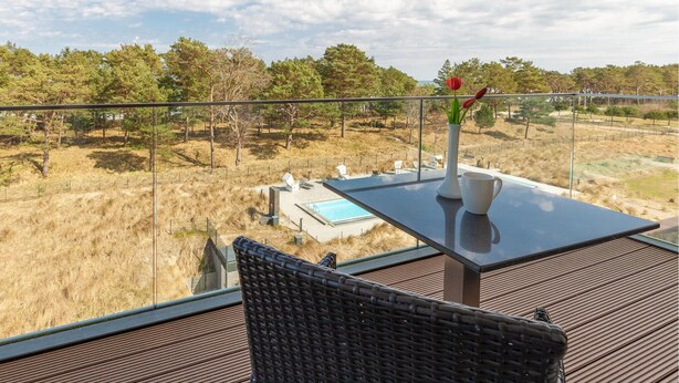 A balcony with a view of a pool and trees, a table with a vase and a cup, and a wicker chair.