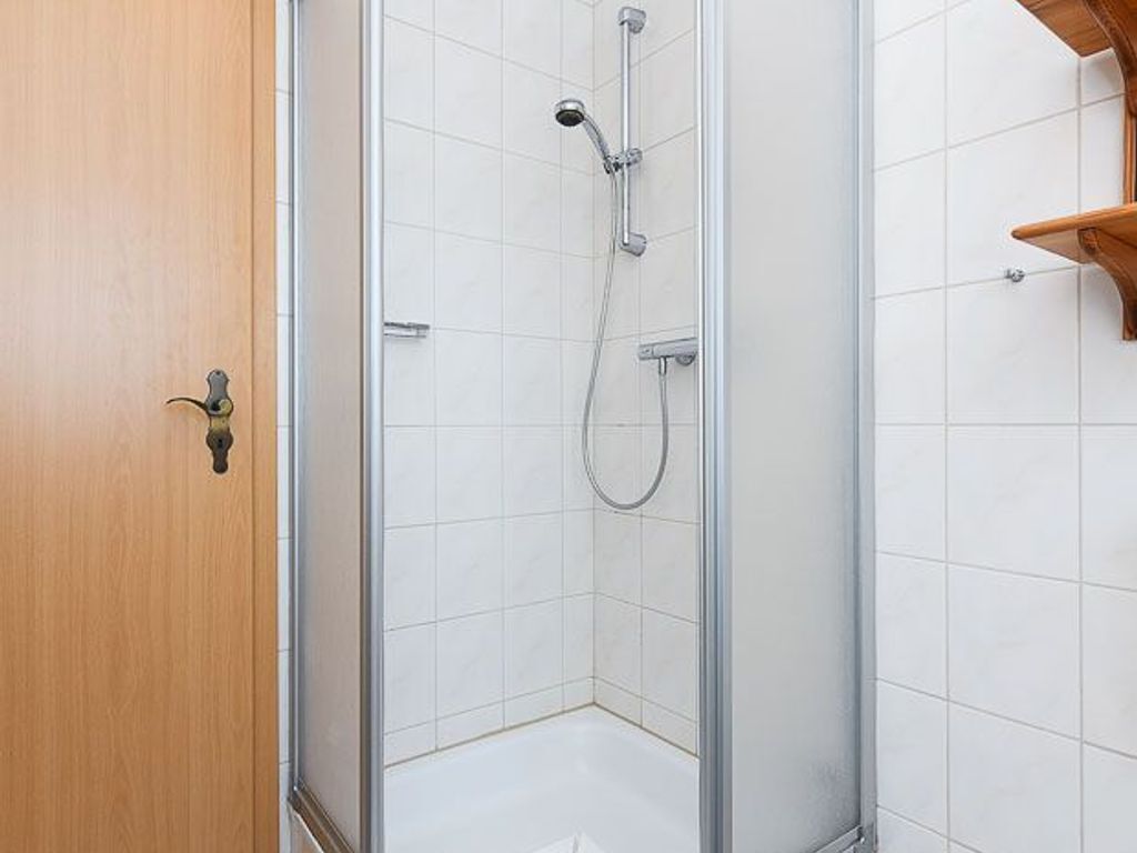 Una doccia con porta in vetro, una porta in legno a sinistra e pareti rivestite di piastrelle bianche.