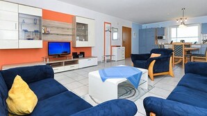 A modern living room with blue sofas, a flat-screen TV, and a kitchen area in the background. - Apartment Achterloog (Werdum)