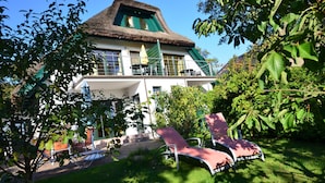 A large house with a thatched roof, a balcony, and a garden with lounge chairs.
