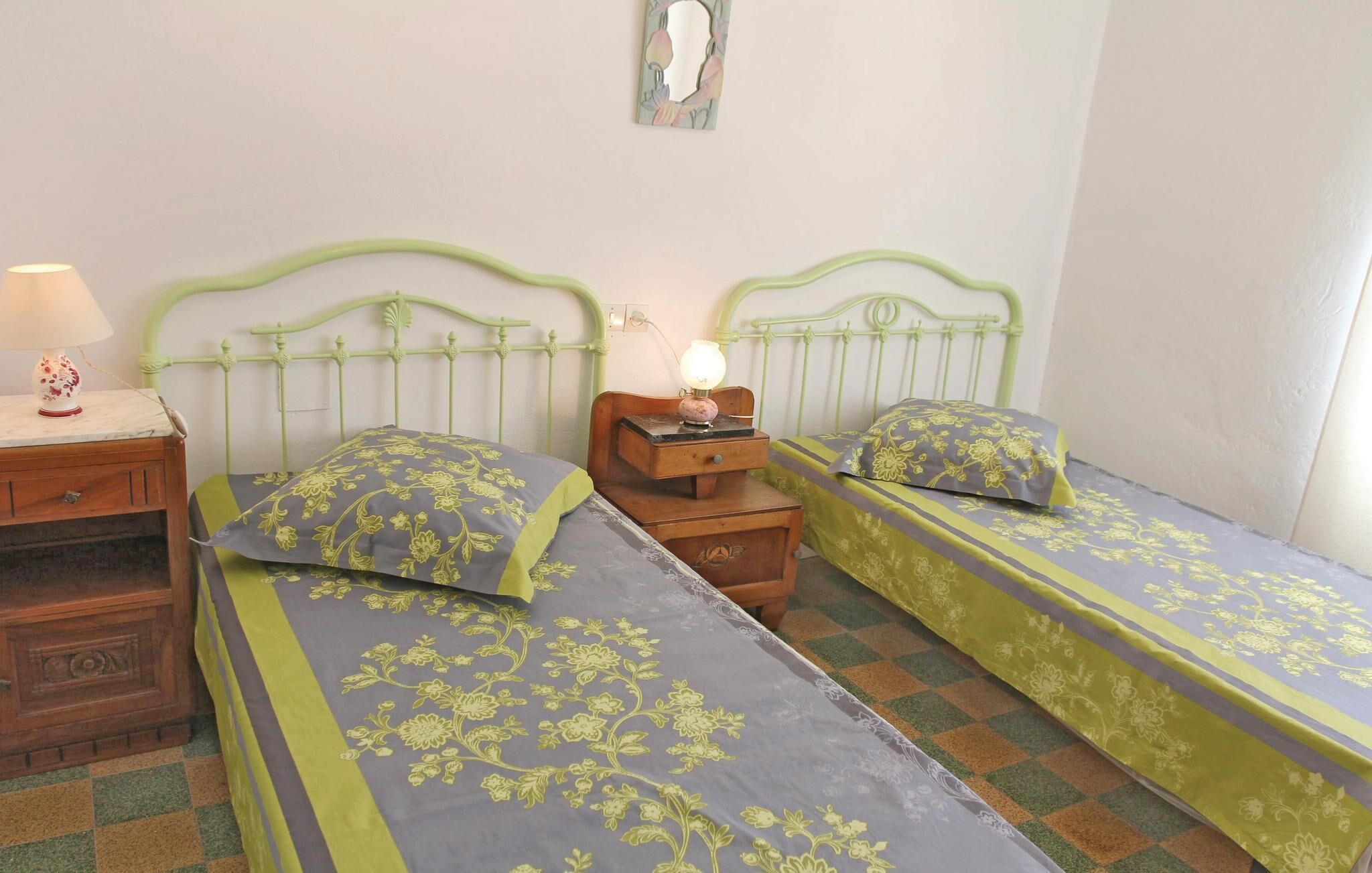 A room with two single beds, each with floral-patterned bedding, a nightstand with a lamp, and a small wooden chest.