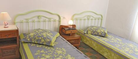 A room with two single beds, each with floral-patterned bedding, a nightstand with a lamp, and a small wooden chest.