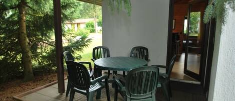 A covered patio with a round table and four chairs, surrounded by trees and a white wall.