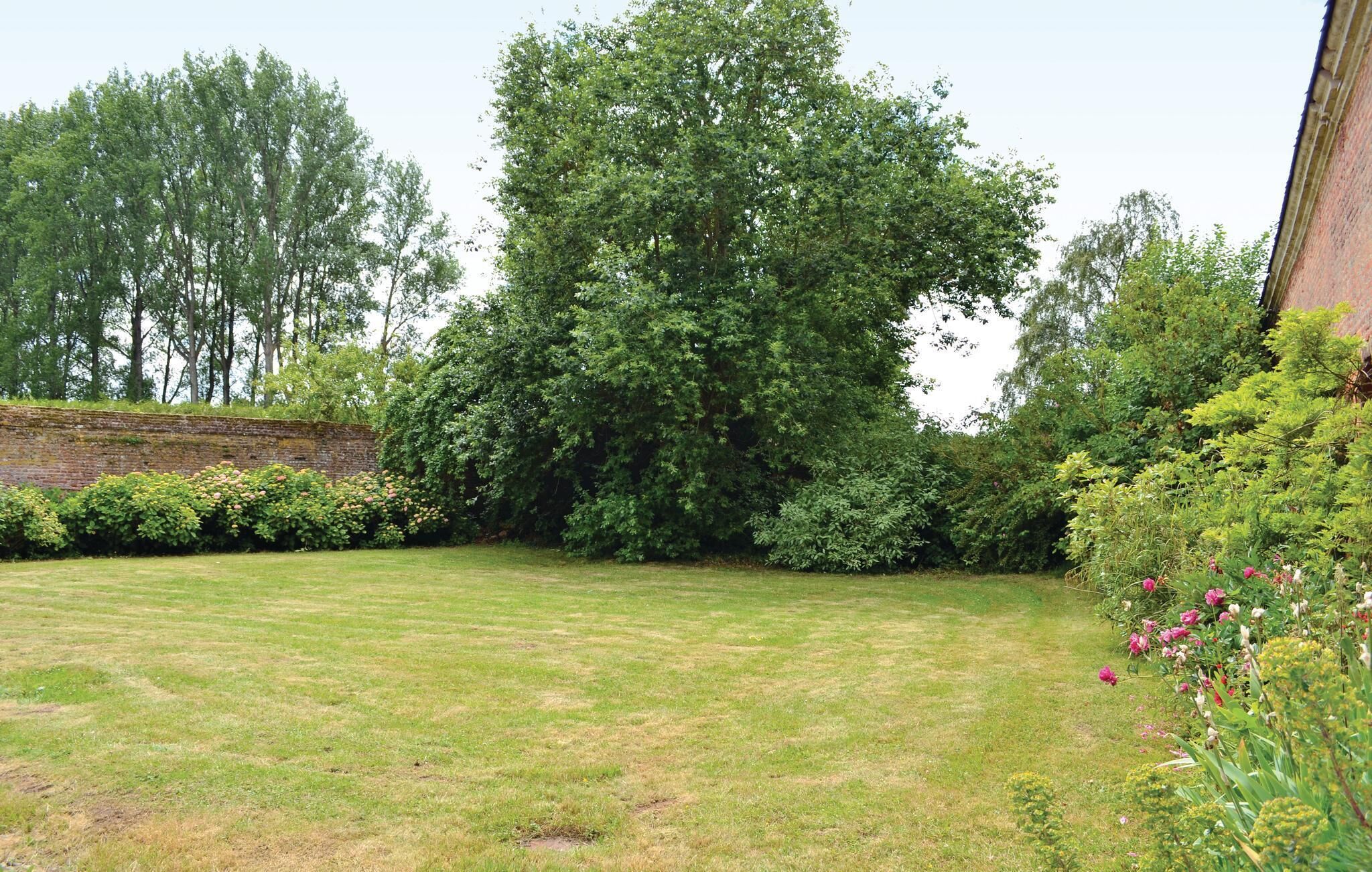 A well-maintained lawn with a variety of shrubs and flowering plants, a brick wall, and tall trees in the background.