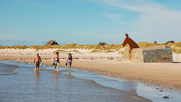 Una playa con gente caminando por la orilla, una estatua de un caballo y cabañas de dunas al fondo.