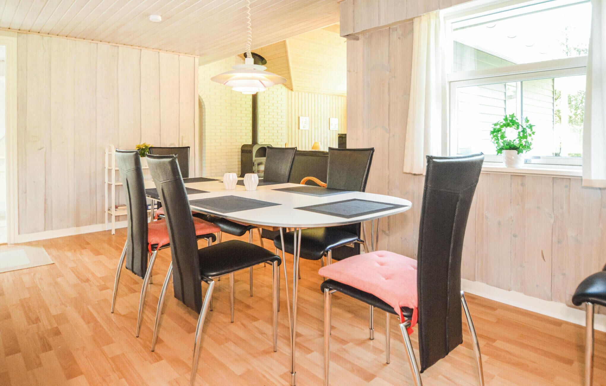 A dining area with a round table, black chairs with red cushions, a hanging light fixture, and a window with a view of greenery.