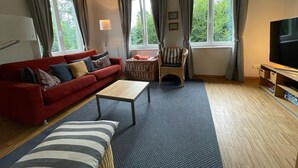 Una sala de estar con un sofá rojo, una mesa de centro de madera, un cojín a rayas, una silla de mimbre y un televisor sobre un mueble de madera.