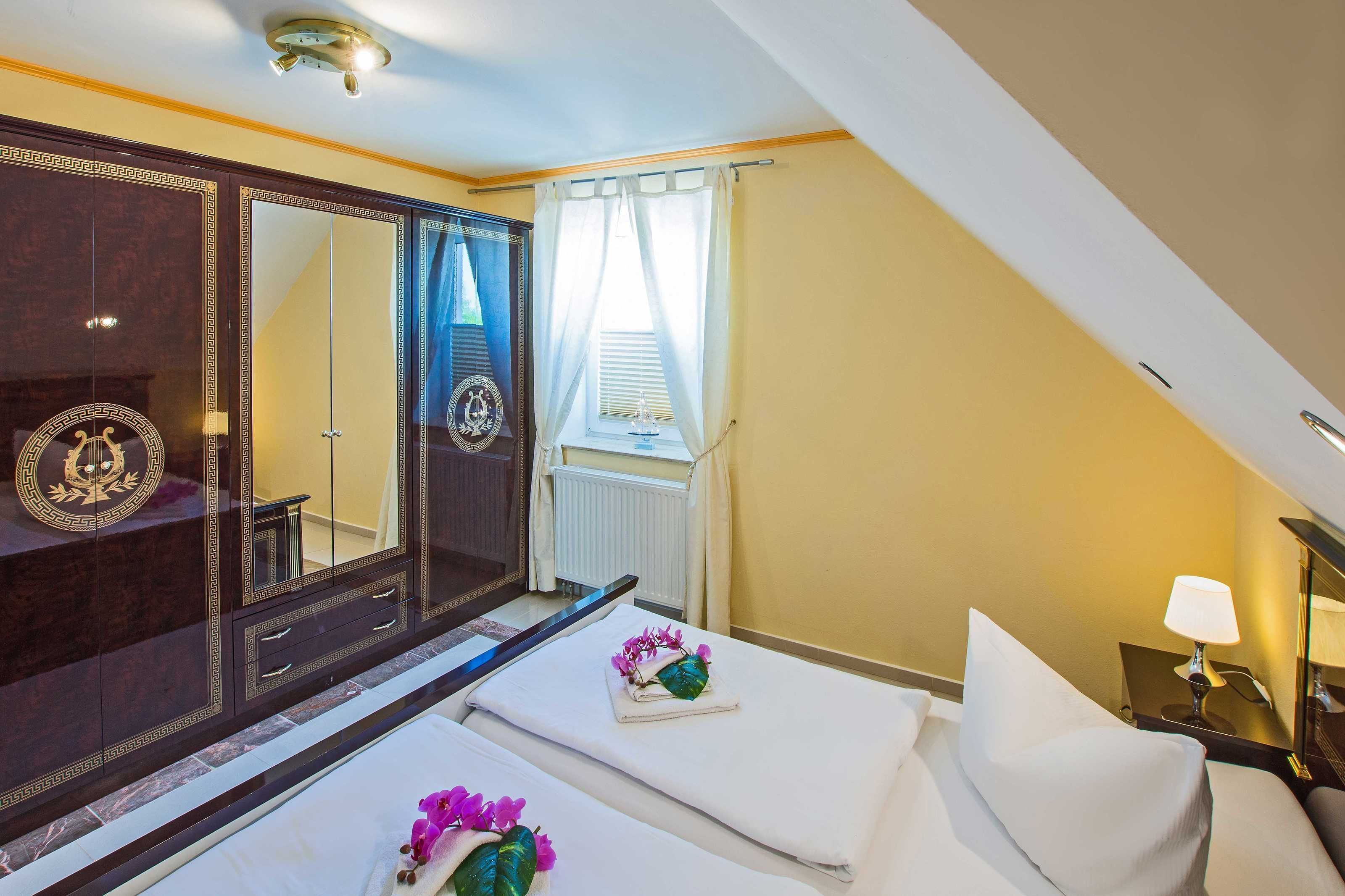 A hotel room with a large dark wooden wardrobe, a bed with white linens, a small table with a lamp, and a window with curtains.