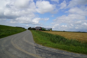Une route de campagne sinueuse traverse un champ, avec une maison et une grange visibles au loin.