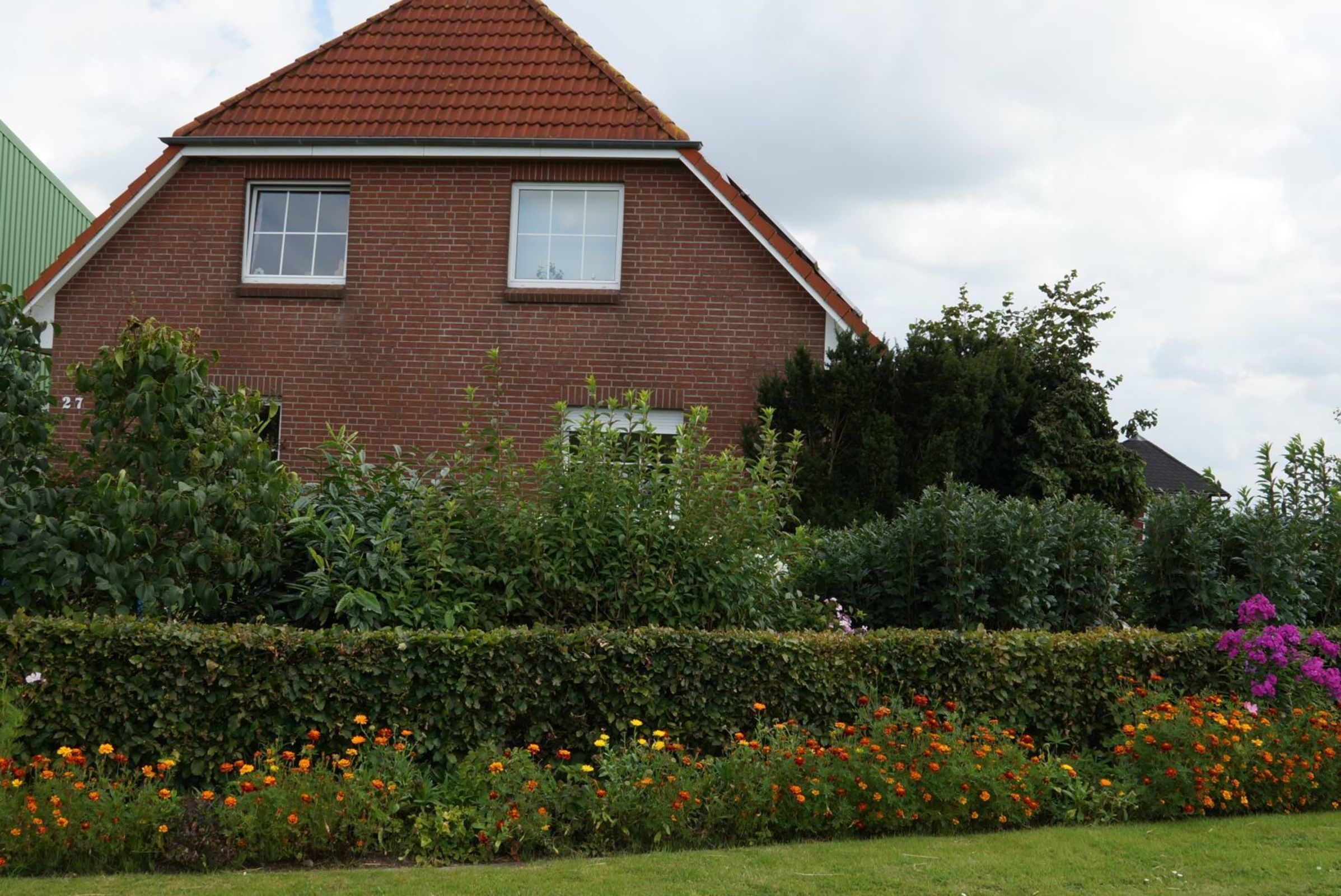 A two-story brick house with a red-tiled roof, two windows, and a well-maintained garden with various flowers and shrubs.