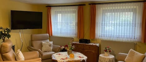 Una sala de estar con sofá, sillón y una mesa de centro. Hay un televisor montado en la pared y una ventana con cortinas.