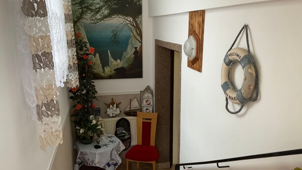 Un comedor con una mesa preparada para dos personas, una silla, una planta decorativa y un cuadro con una escena costera.