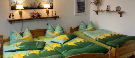 Un dormitorio con dos camas de madera, ropa de cama estampada con motivos florales en verde, un cuadro sobre las camas, un estante con objetos decorativos y una fotografía enmarcada en la pared.