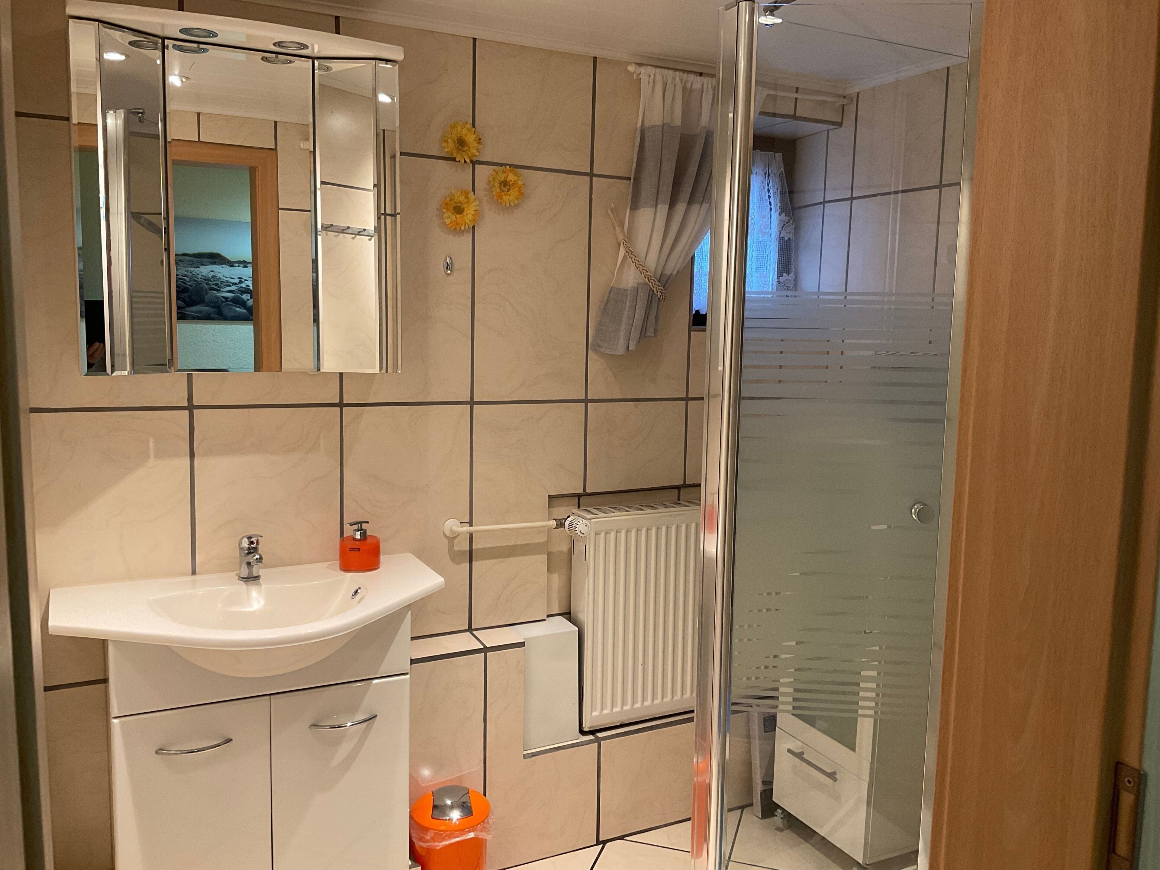 Een badkamer met een wit wastafel, spiegel en betegelde muren. Er is een douchegedeelte met een glazen deur en een handdoekhouder.