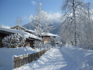 Ein verschneiter Pfad führt zu einem Holzbauten mit schneebedecktem Dach, umgeben von Bäumen und Bergen.