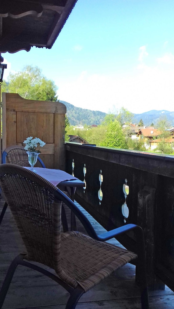 A balcony with wicker chairs, a table with a vase of flowers, and a view of a mountainous landscape.