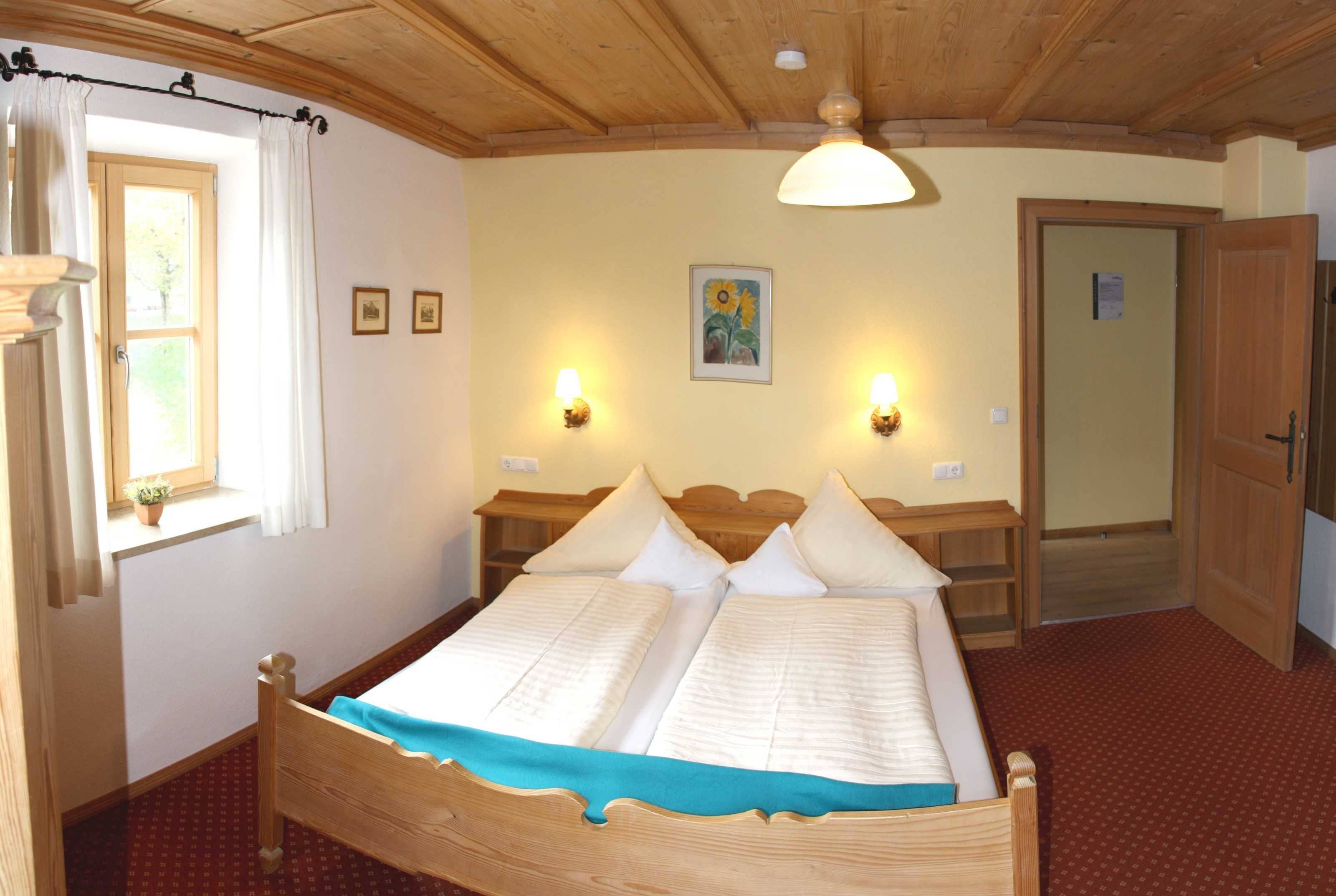 A double bed with a wooden frame, two bedside tables, a window with curtains, a wall-mounted light fixture, and a door.