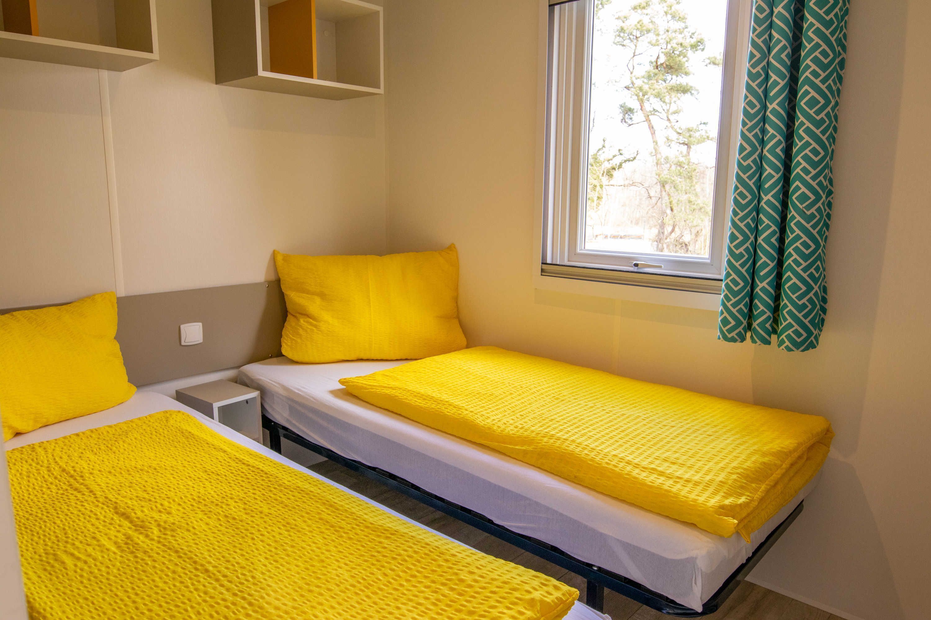 Una habitación con dos camas, cada una con una colcha y almohadas amarillas, una ventana con cortinas y un estante sobre las camas.