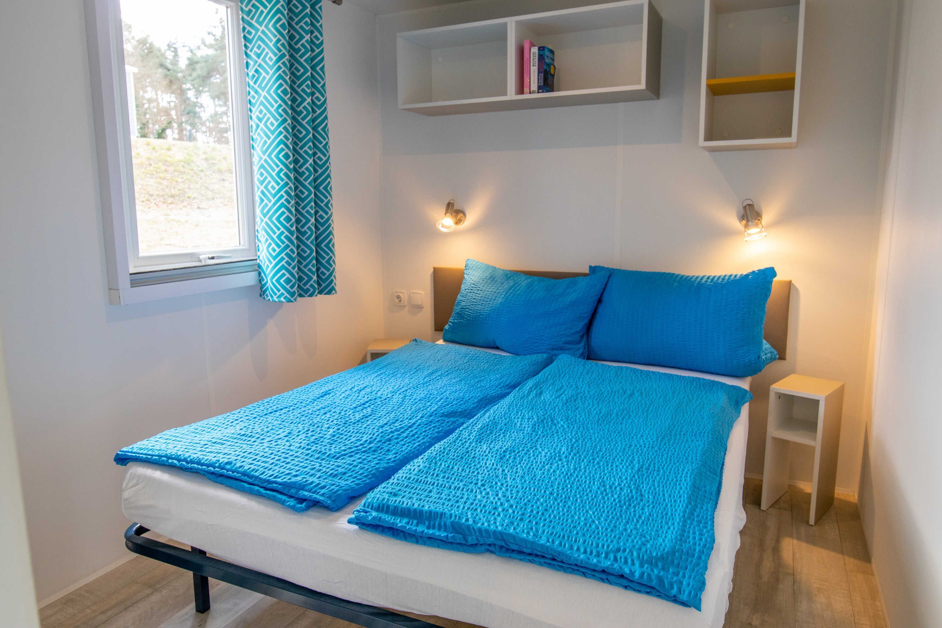 A bed with a white mattress and blue bedding, a window with a geometric curtain, and two bedside tables with wall-mounted lights.
